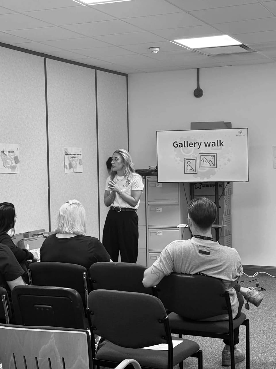 A presenter standing beside a wall screen headed 'Gallery walk', with flipchart sheets and sticky notes on the wall behind and a seated group in front.
